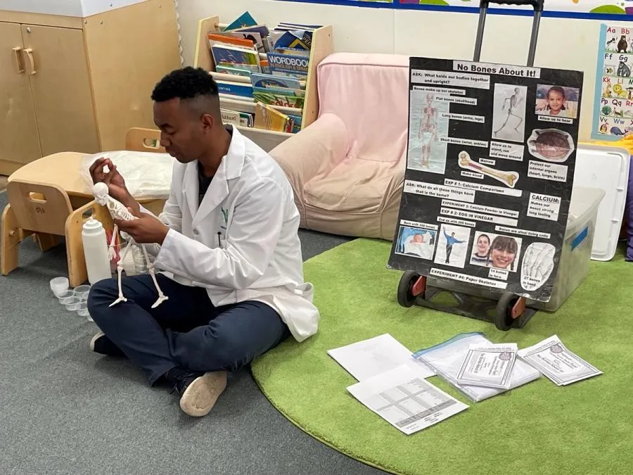 An adult man in a lab coat sits on the floor in front of a poster about bones. He is holding a model skeleton.