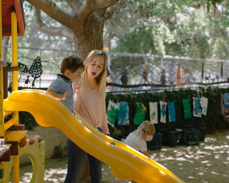 A blonde woman helps a young child slide down a yellow slide.