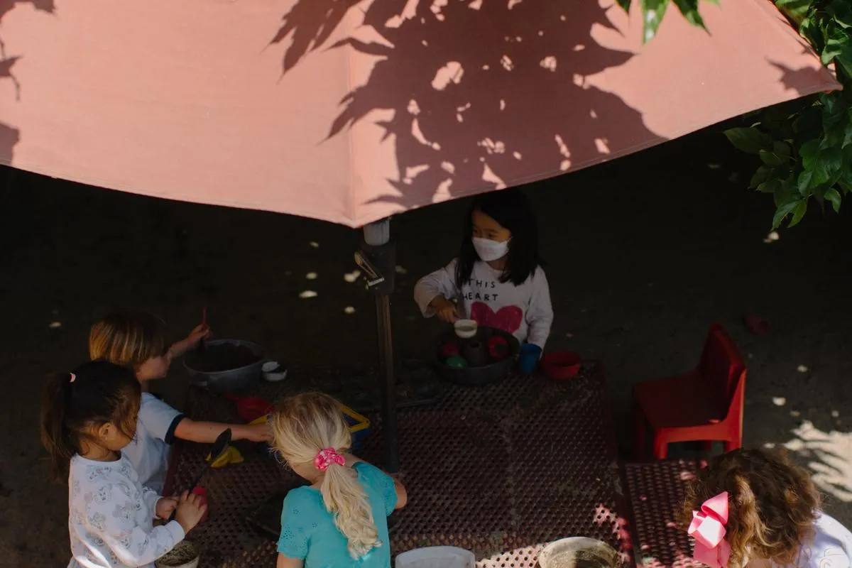 Small children wear masks and gather around a table under a red umbrella and surrounding trees.