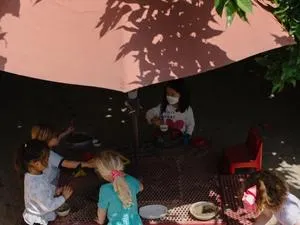 Small children wear masks and gather around a table under a red umbrella and surrounding trees.