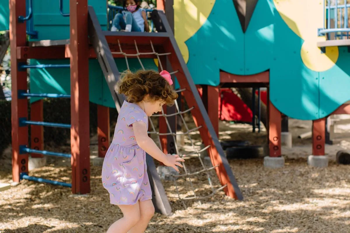 A small child in a purple dress jumps off a log in front of playground equipment.