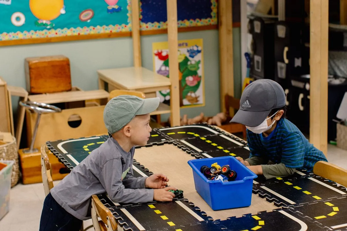 Two children wearing baseball hats lean on a low table with a road mat and a box of cars.
