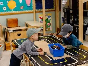 Two children wearing baseball hats lean on a low table with a road mat and a box of cars.