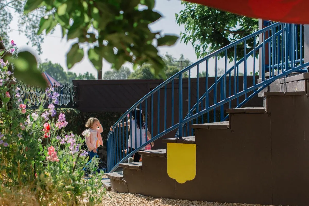 Two small children play on a playground structure with large stairs with a blue railing next to a flower bed.