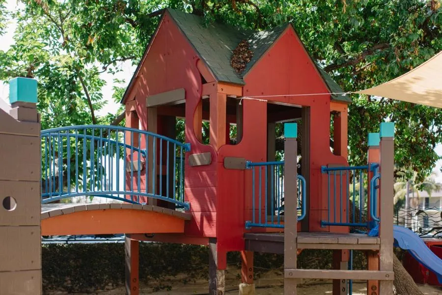 View of a playground structure. A red house with a bridge and blue slide under a large tree.