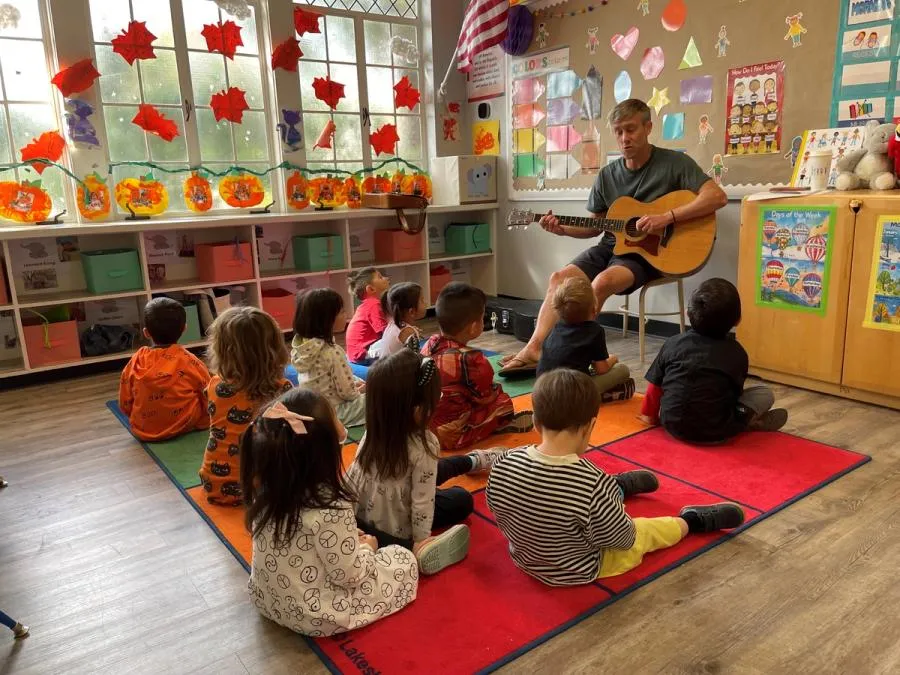 An adult man sits on a chair with a guitar in front of eleven children siting on a red rug. They are in a class room decorated with cutouts of fall leaves.