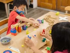 Two young children wearing masks play with wooden toys at a table.