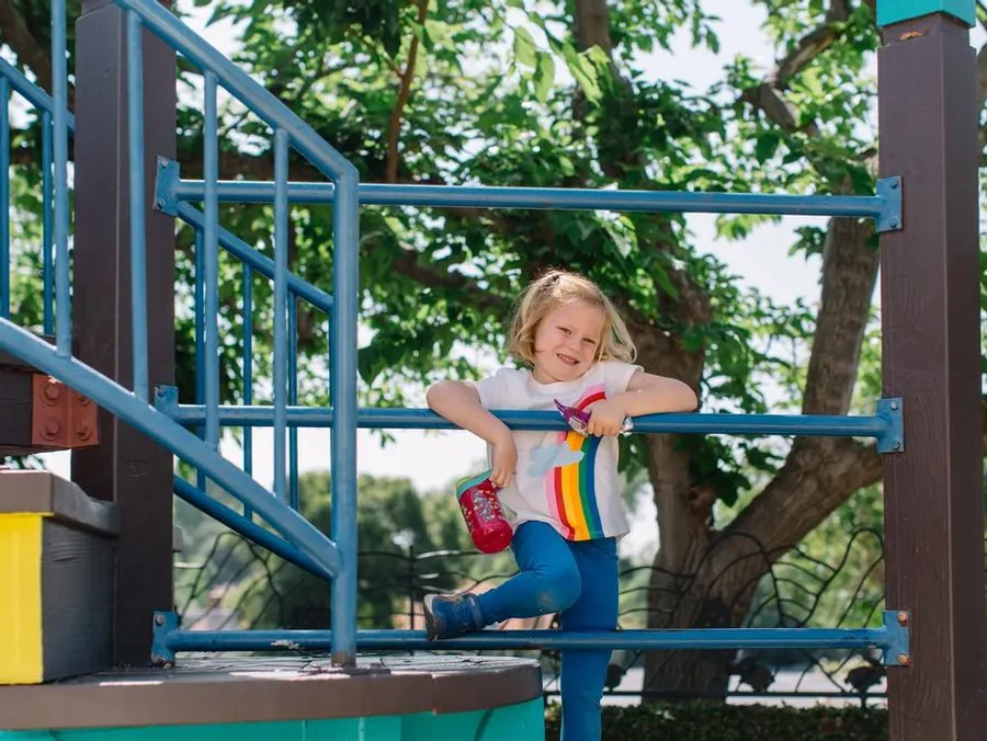 A child wearing a rainbow shirt smiles and hangs on the blue railing of a playground structure.