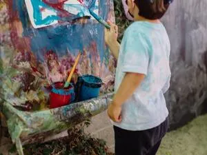From behind: a young child with a baseball hat paints red and blue lines on an easel outside.