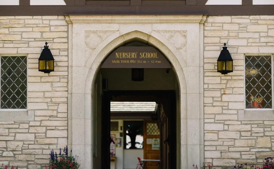 A flat stone building with a center stone arch. A sign hangs from the arch and says: Nursery School.
